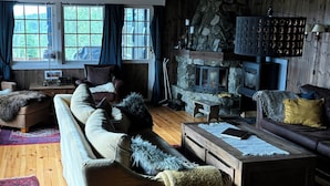 A cozy living room with a stone fireplace, wooden floors, and a large window with curtains.