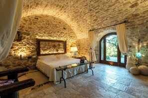 A stone-walled bedroom with a bed, bedside table, lamp, and a view through an arched window. - Medieval tower in Sibillini Mountains Park (Montefortino FM)