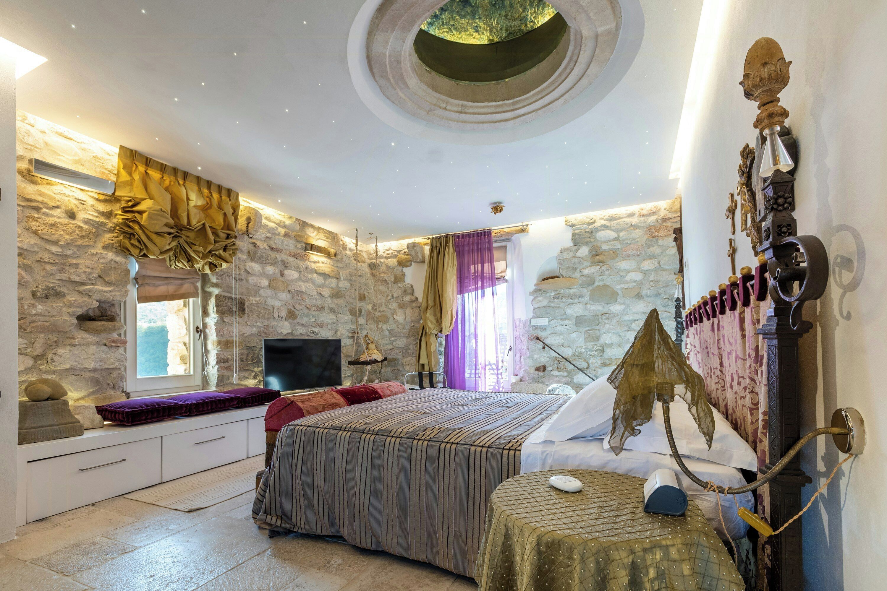 Une chambre avec des murs en pierre, un lit, une télévision et un plafond au design original.