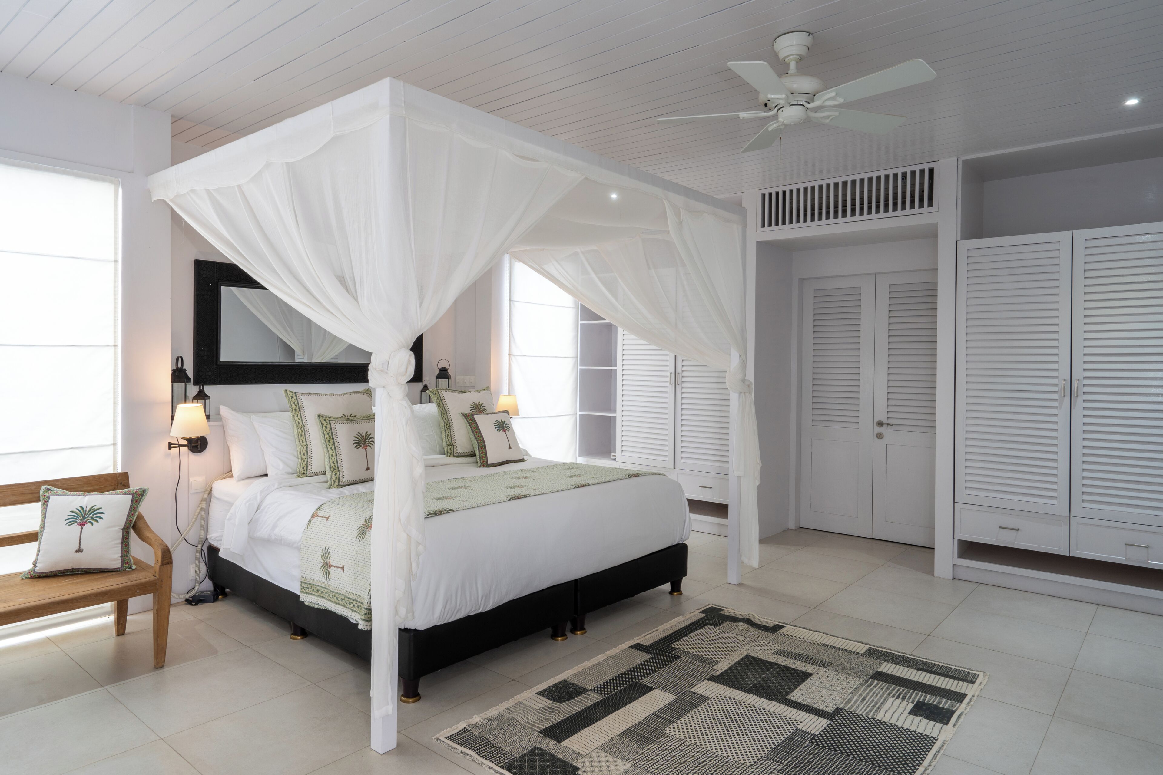 A four-poster canopy bed with a white bedspread and pillows, a wooden bench, a patterned rug, and a ceiling fan.