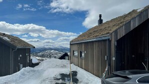 Et snøklædt udendørs område med træhuse, en bil og et bjerglandskab i baggrunden.