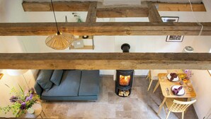 Ein Loft-Zimmer mit freiliegenden Holzbalken, einer Couch, einem Esstisch für zwei Personen und einem Kaminofen.