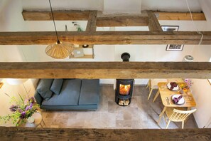 Ein Loft-Zimmer mit freiliegenden Holzbalken, einer Couch, einem Esstisch für zwei Personen und einem Kaminofen.