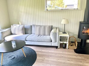 Ein gemütliches Wohnzimmer mit grauem Sofa, einem kleinen runden Tisch, einer Lampe, einem Korbwäschekorb und einem Kaminofen.