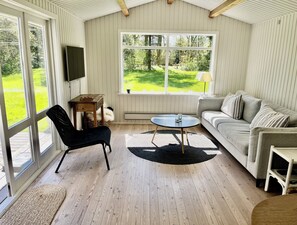 Ein Zimmer mit Holzboden, einer Couch, einem Sessel, einem Fernseher und einem großen Fenster mit Blick auf Grünflächen.