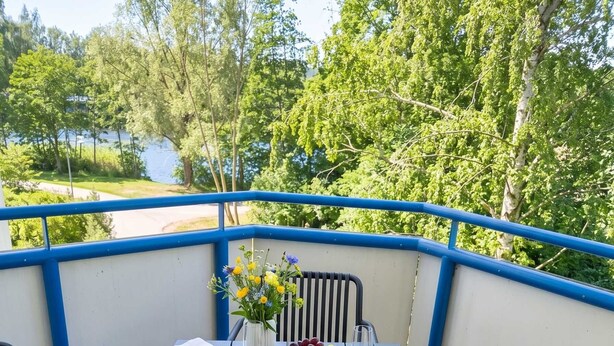 A table set for two with plates, cutlery, glasses, and a bowl of fruit, surrounded by chairs, with a view of trees and a body of water. - Moewennest in Rostock (Rostock)