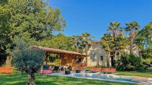 A covered outdoor seating area with tables and chairs, surrounded by lush greenery and palm trees. - American-style villa with pool in Montalivet (Vendays-Montalivet)