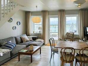 A living room with a sofa, coffee table, dining table, chairs, and a view of the sea through large windows. - Holiday home with harbour view in Klintholm (Borre)