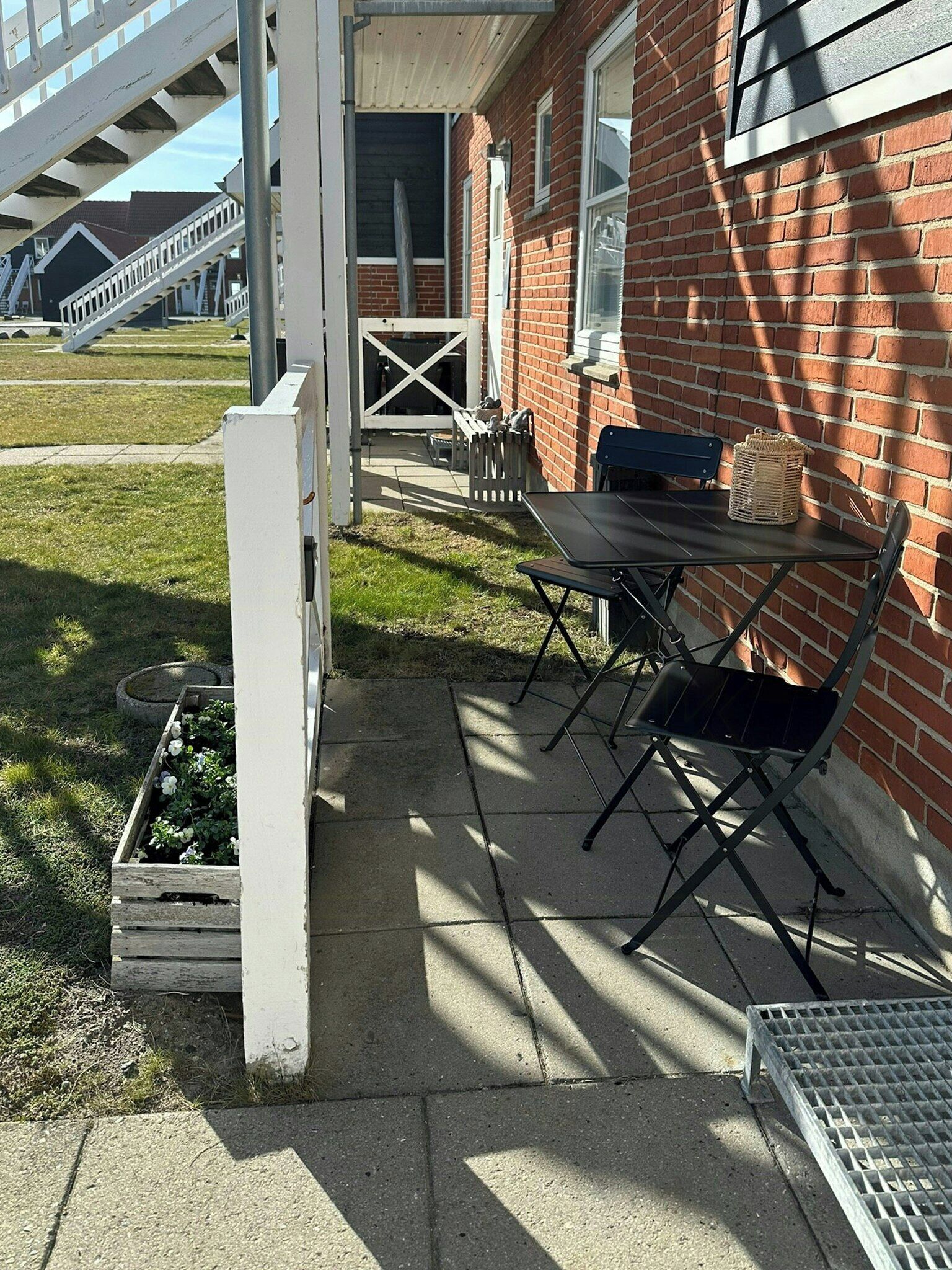 A patio with a table and chairs, a white fence, and a brick building in the background.