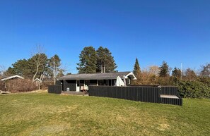 A single-story building with a covered patio area, a fenced yard, and a clear blue sky. - Cozy summer house at Odden (Sjællands Odde)