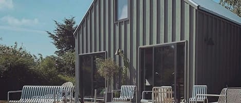 A modern building with a sloped roof and large windows, a wooden deck with metal chairs, and a small tree in the center.