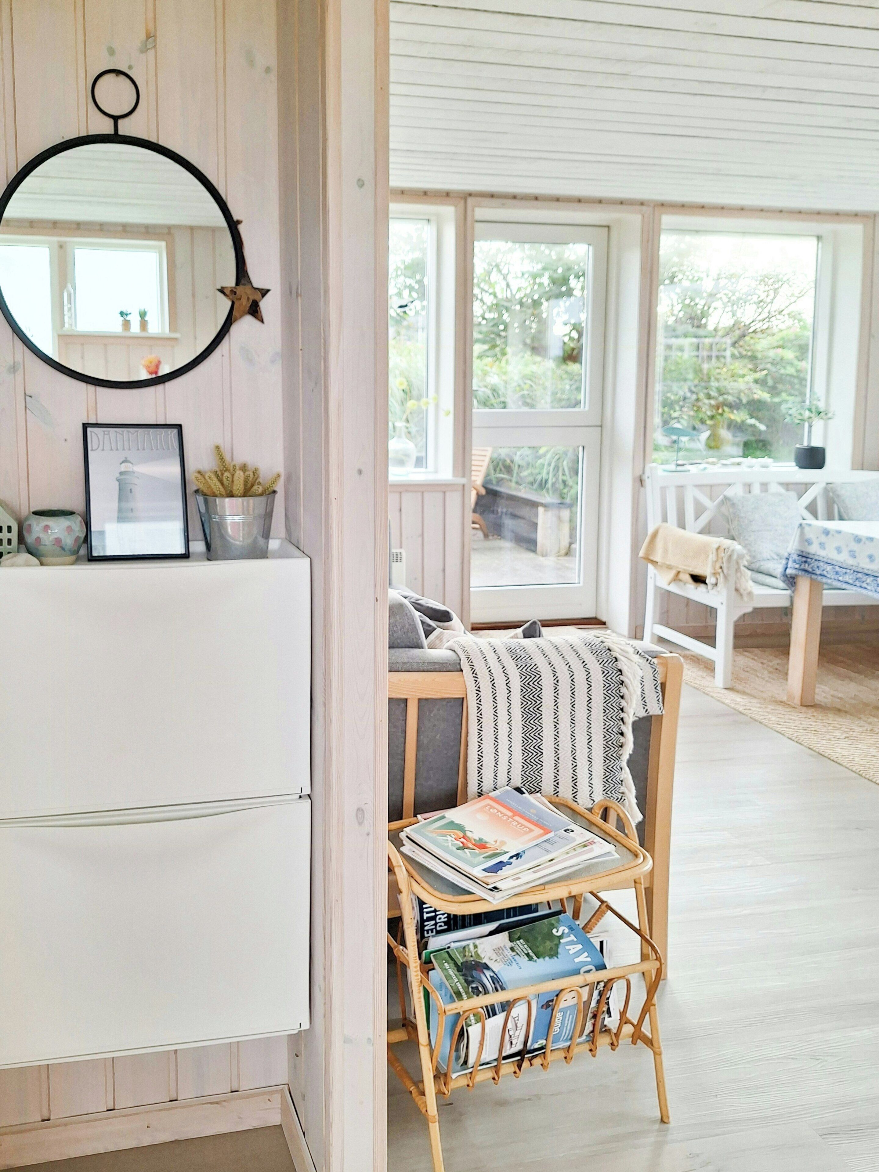 A room with a white wardrobe, a framed picture, a metal bucket, a wicker chair with magazines, and a view of the outdoors through large windows.