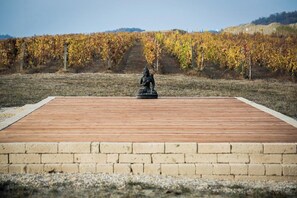 Eine Statue auf einer Holzplattform, im Vordergrund eine Backsteinmauer und im Hintergrund Rebenzeilen.