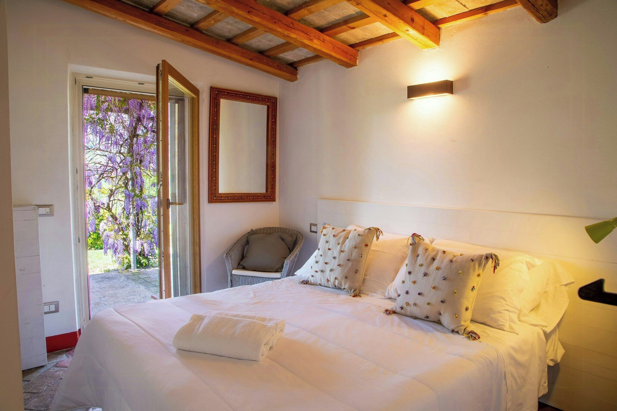 Une chambre à coucher comprenant un lit, une chaise, un miroir et une porte donnant sur un espace extérieur agrémenté de fleurs violettes.