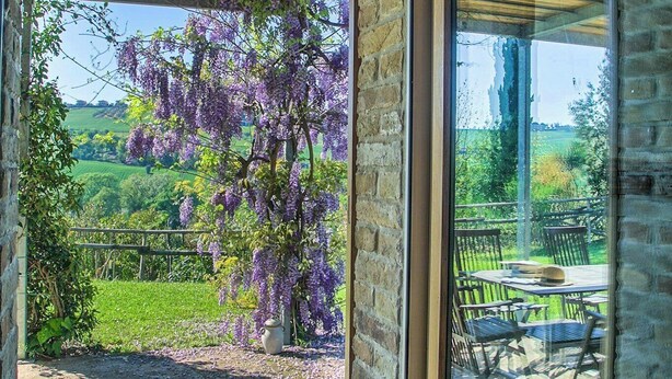 En glasdør fører ud til en patio med udsigt over en frodig have og et lilla blomstrende træ.