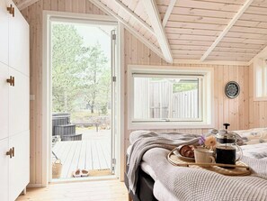 A wooden cabin bedroom with a bed, a window, and a view of a deck and trees. - House in the heather with sea view on Odden (Sjællands Odde)