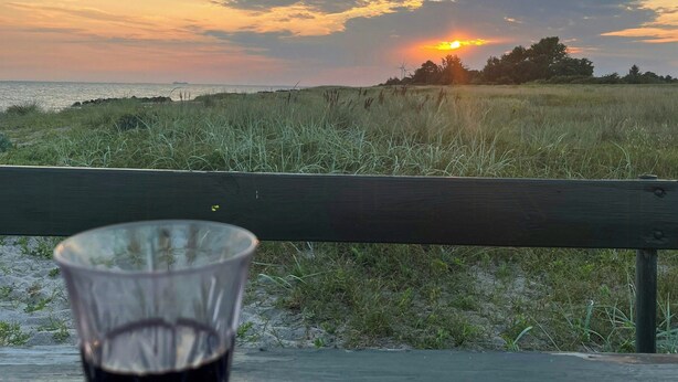 Um copo de vinho tinto sobre um banco de madeira com vista para uma praia ao pôr do sol.