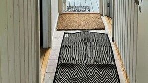 Um corredor com piso de madeira, um tapete com padrão preto e branco, uma porta de madeira e um espelho fixado na parede.