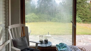 Ein Zimmer mit Blick auf eine grüne Fläche, einem Korbwägelchen, einem Sessel mit Kissen und einem kleinen Tisch mit Gläsern.