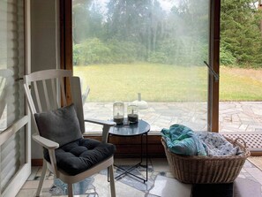 Ein Zimmer mit Blick auf eine grüne Fläche, einem Korbwägelchen, einem Sessel mit Kissen und einem kleinen Tisch mit Gläsern.