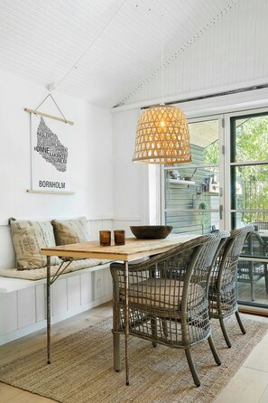 Ein modernes Interieur mit weißer Decke, einem Holztisch mit Schale und Gläsern, Weidekorbstühlen und einer hängenden Leuchte.