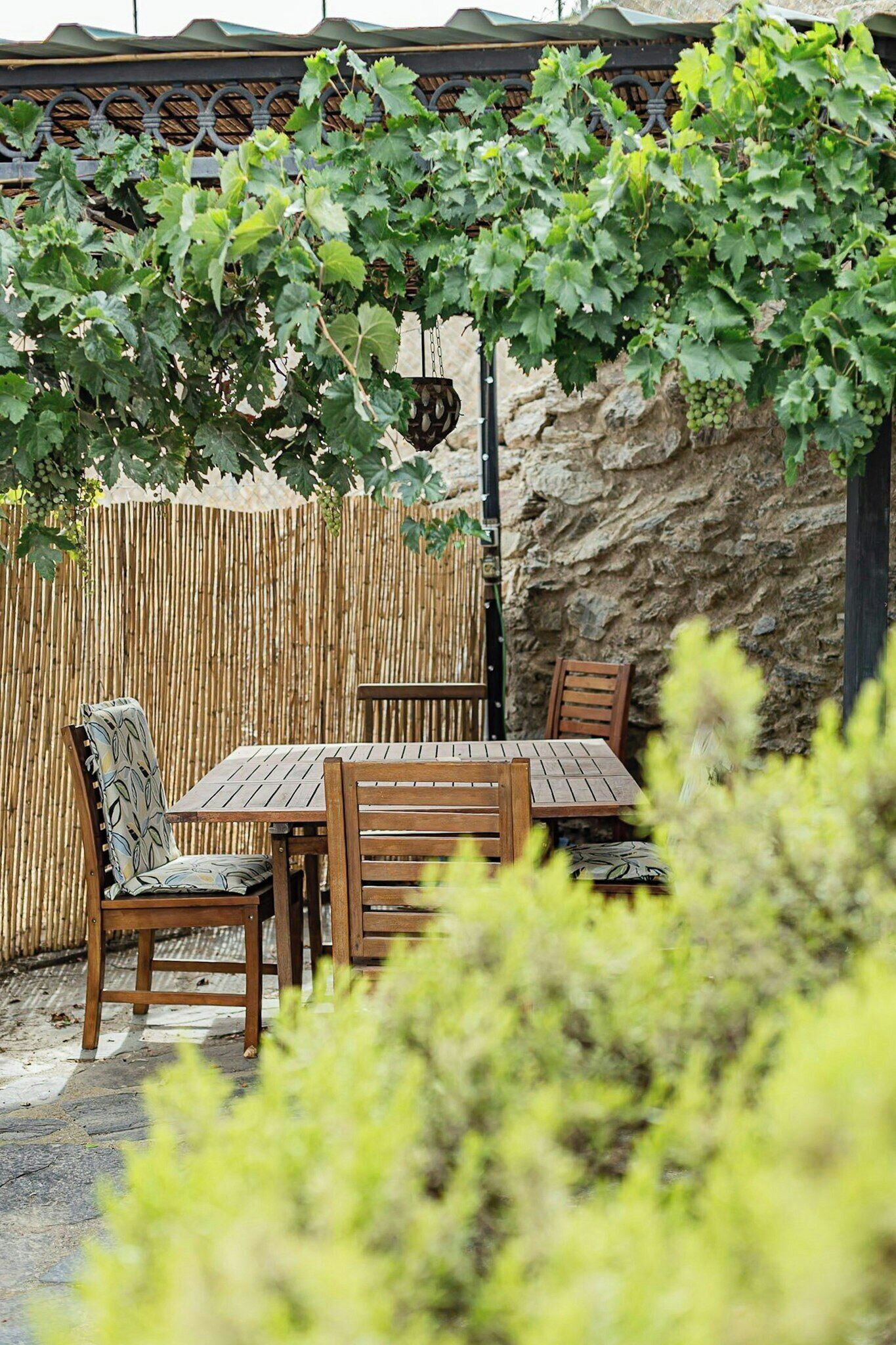 A wooden table and chairs set outdoors, with a stone wall and a bamboo fence in the background.