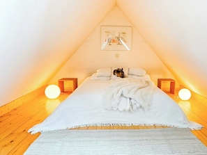 Ein schrägwandiges Schlafzimmer im Dachgeschoss mit einem Bett, einer weißen Decke, zwei Nachttischlampen und einem gerahmten Bild an der Wand.