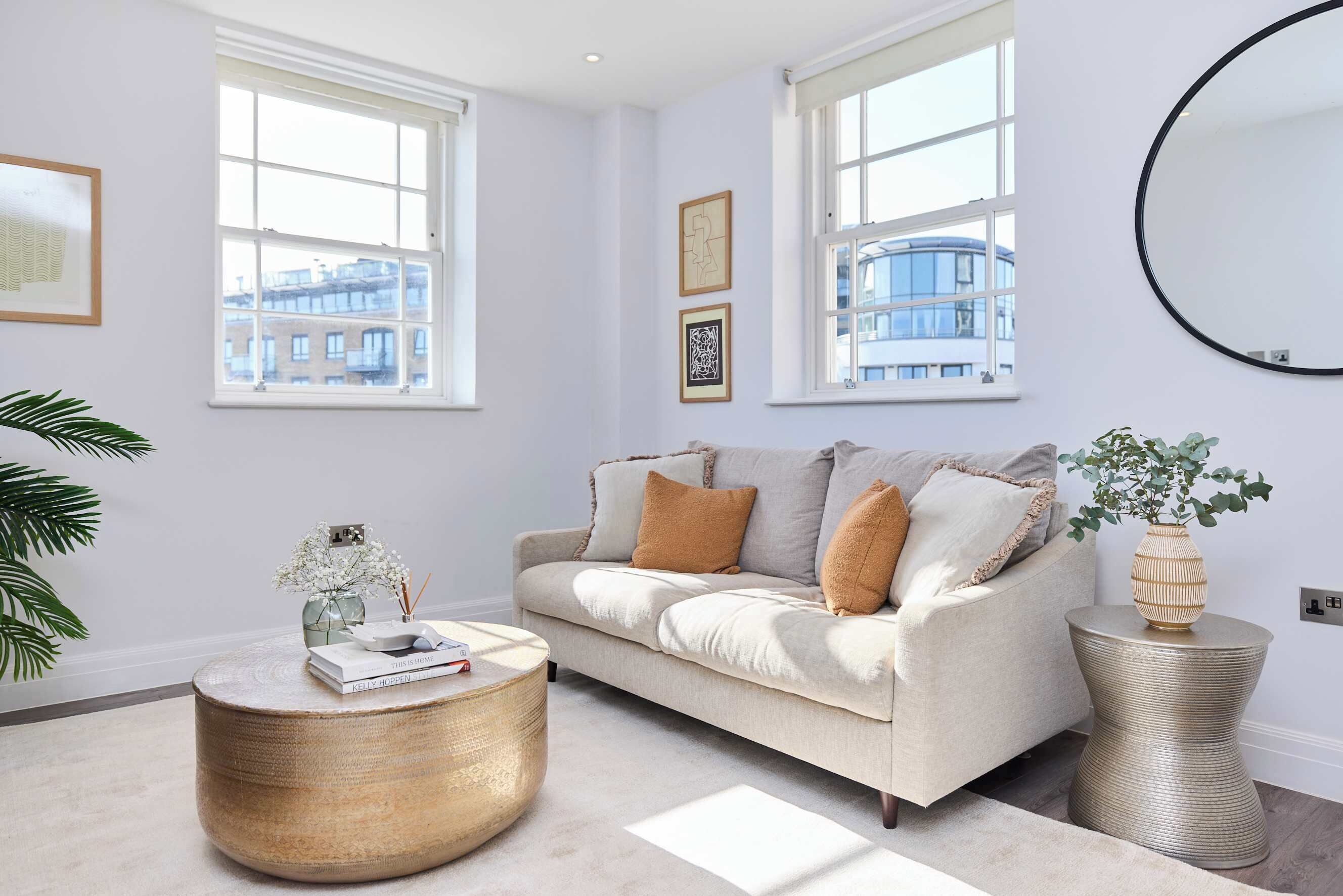 A living room with a beige sofa, round coffee table, and side table with a vase, large windows, and a mirror on the wall.