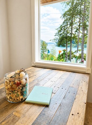 A wooden floor, a glass jar with seashells, and a book titled 'Guest Book Be Our Guest' near a window with a view of trees and water.