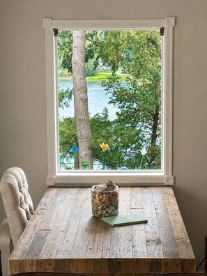 A wooden table with a jar of seashells and a green book, a chair, and a window with a view of a lake and trees.