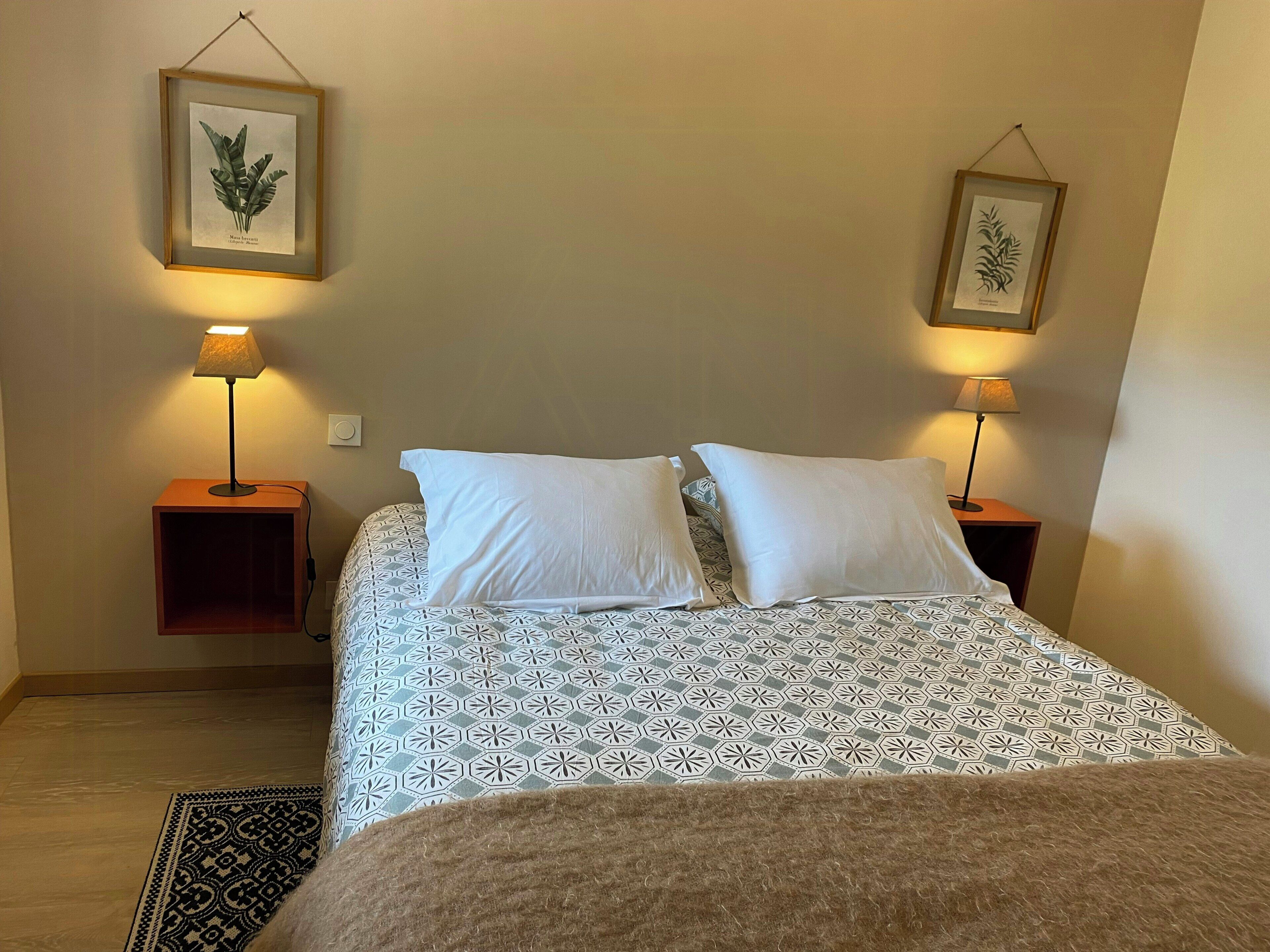 A bedroom with a bed, two bedside tables, two lamps, and two framed botanical prints on the wall.