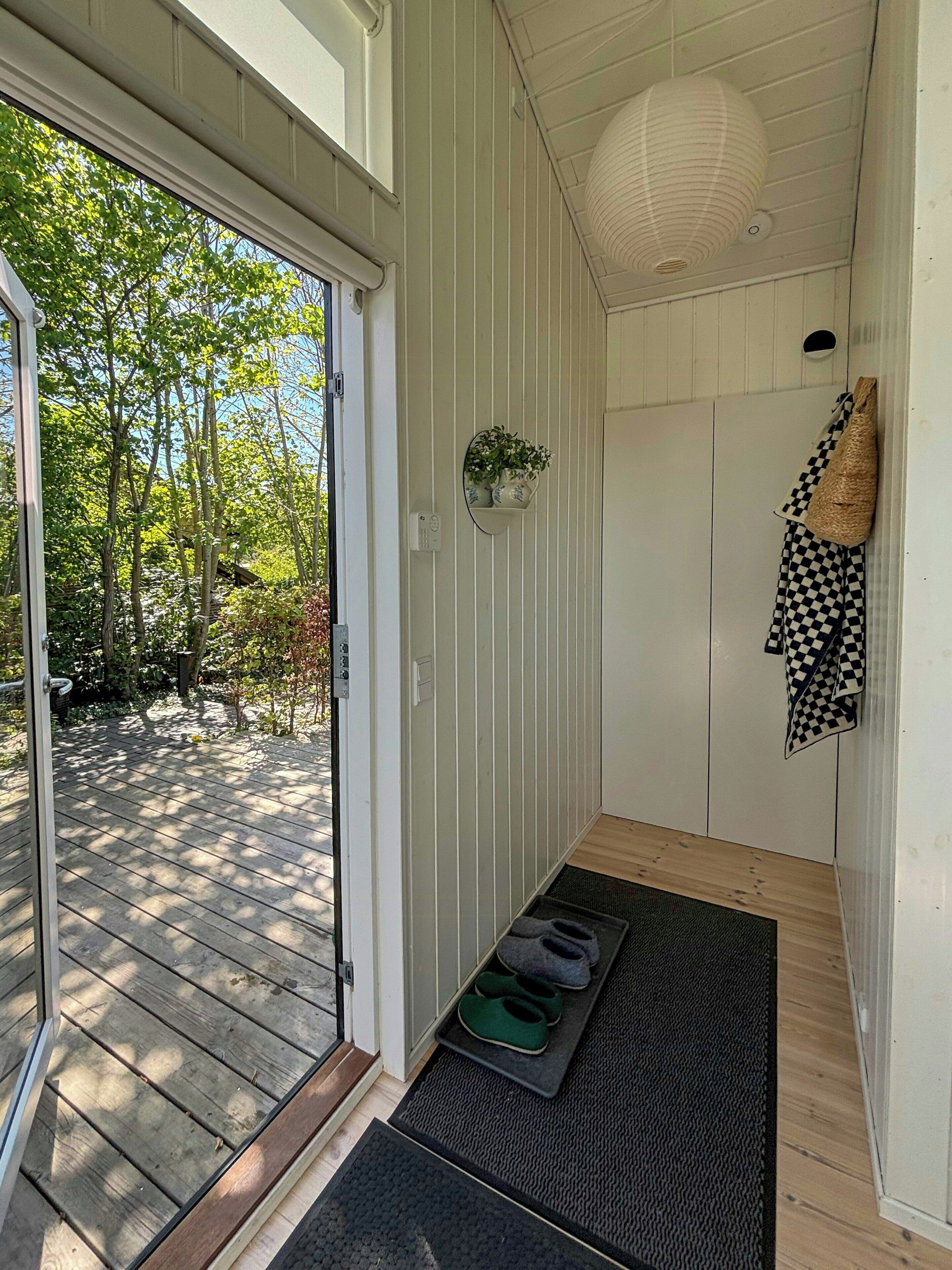 A wooden deck leading to a room with a sliding door, a small shelf with plants, a checkered towel, and a woven bag.