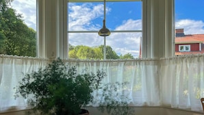 Ett rum med ett stort fönster, en krukväxt, ett bord med böcker och en hängande lampa.