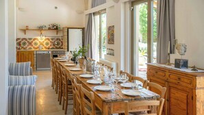Ein Essbereich mit einem langen Holztisch, gedeckt für eine Mahlzeit, Holzsesseln, einer Küche im Hintergrund und Blick ins Freie durch große Fenster.