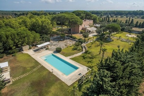Un'ampia piscina rettangolare circondata da un solarium in pietra, con un'area verde e alberi sullo sfondo.