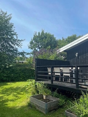 A wooden deck with a table and chairs, surrounded by greenery and a building with a sloped roof. - Summer house with sunset over Sejerø Bay (Asnæs)
