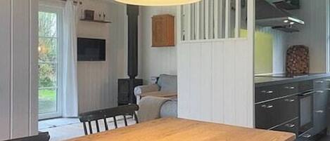 A dining area with a wooden table and chairs, a hanging light fixture, and a view into the kitchen.