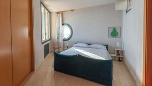 Um quarto com cama, janela, cadeira, mesa de cabeceira e prateleira na parede.