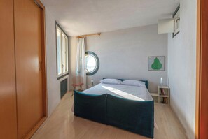 Ein Schlafzimmer mit Bett, Fenster, Stuhl, Nachttisch und einer an der Wand befestigten Ablagefläche.