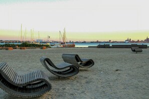 A beach with lounge chairs and a view of the water and distant cityscape. - Family home near beach in Altefähr on Rügen (Altefähr)