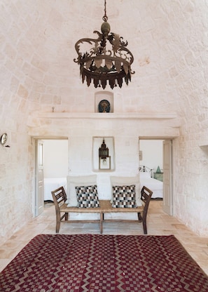 A room with a stone wall, a wooden bench with checkered cushions, a patterned rug, and a hanging chandelier. - Historic trullo in Valle d'Itria (Ostuni)