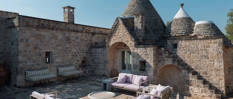 A courtyard with stone walls, a stone building with conical roofs, and outdoor furniture.