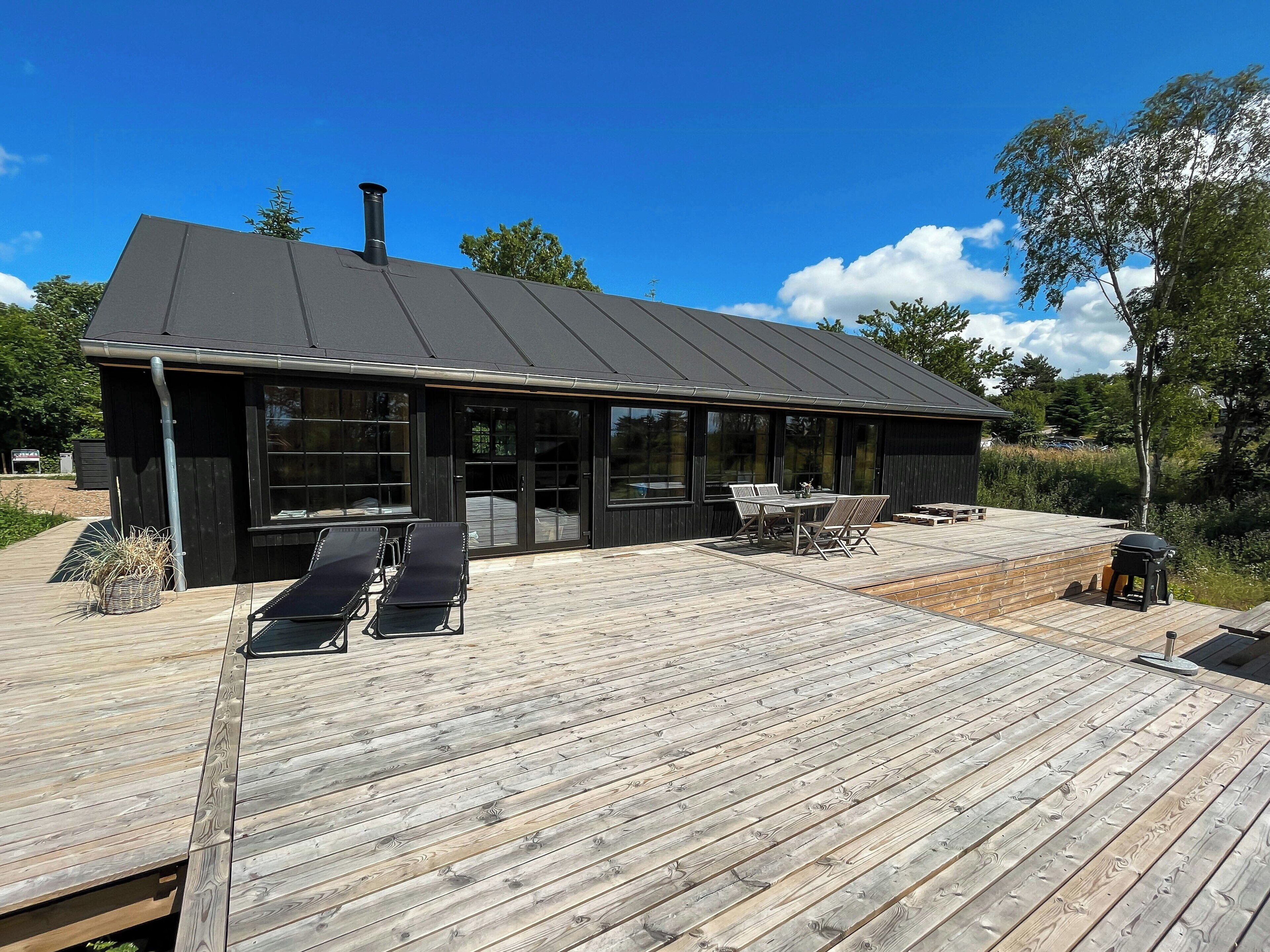 A modern building with a flat roof, large windows, and a wooden deck with lounge chairs and a table.