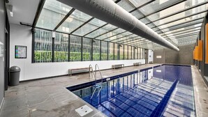 An indoor swimming pool with a glass ceiling, a bench, and a trash bin. - Exclusive Stays - Southbank Grand (Southbank)