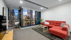 A modern living room with a red sofa, a coffee table, and a TV. There is a large window with a city view. - Exclusive Stays - Southbank Grand (Southbank)