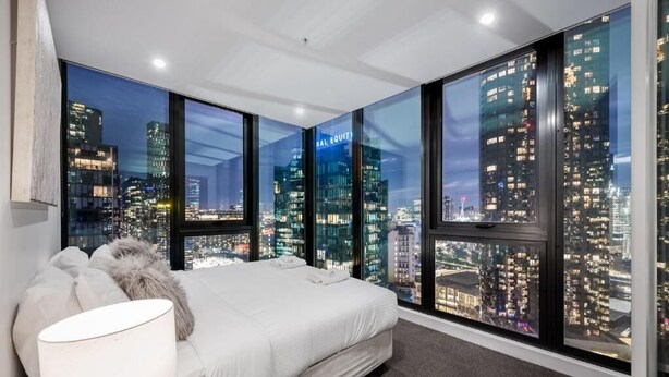 A bedroom with a city view, a bed, a lamp, and a large window. - Exclusive Stays - Southbank Grand (Southbank)