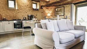 A modern living room with a light-colored sofa, a wooden dining table, a kitchen area with appliances, and a stone wall. - Charming mas with medieval church views (Sainte Croix)