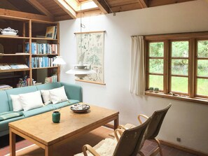 Ein gemütliches Zimmer mit Holzbalkendecke, einem blaugrünen Sofa, einem Holztisch, einem Bücherregal und einem Fenster mit Blick auf Grünflächen.
