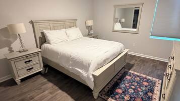 A bedroom with a bed, a nightstand, a lamp, a ceiling fan, a mirror, and a window with blinds.