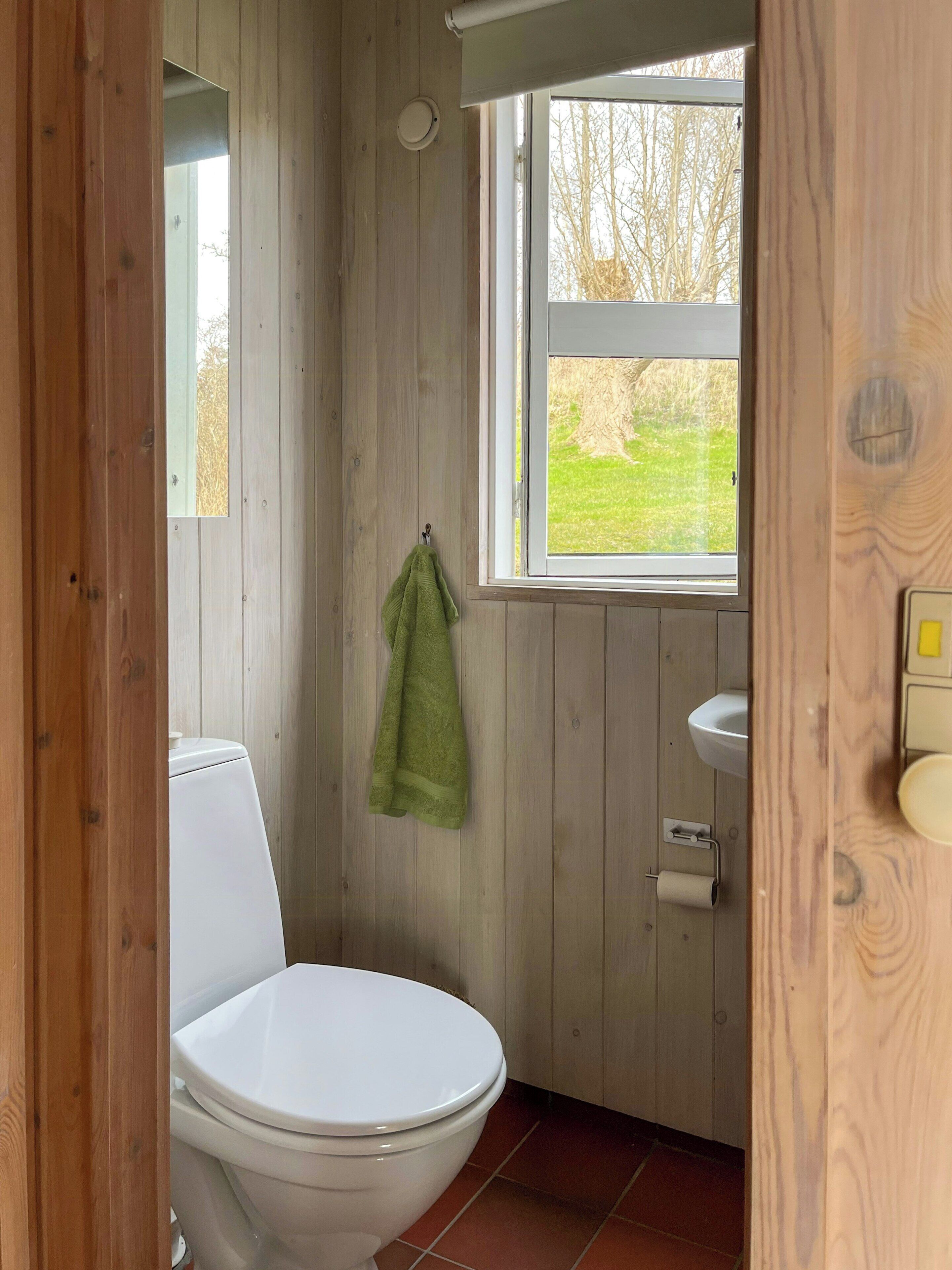 A bathroom with wooden walls, a white toilet, a green towel hanging on the wall, and a window with a view of trees.